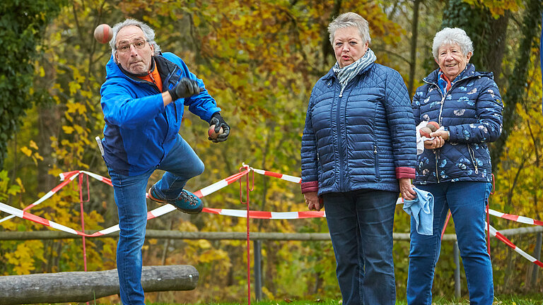 Drei ältere Menschen spielen in einem herbstlichen Park Boggia. Im Vordergrund wirft ein Mann gerade konzentriert eine Kugel, während zwei Frauen seinen Wurf im Hintergrund gespannt beobachten.